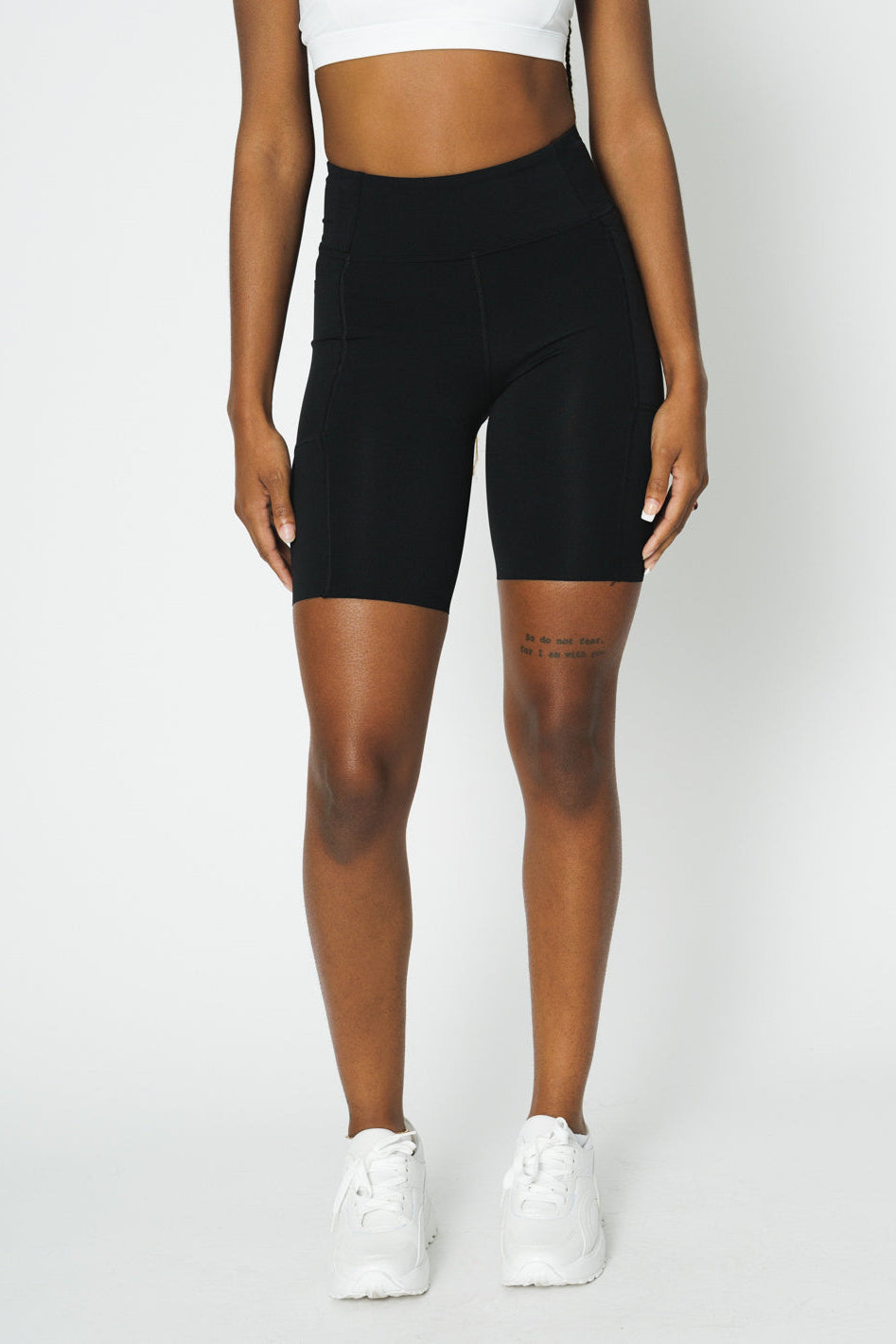 A model wearing black high-rise biker shorts with a raw hem and three pockets, paired with a white top and white sneakers.